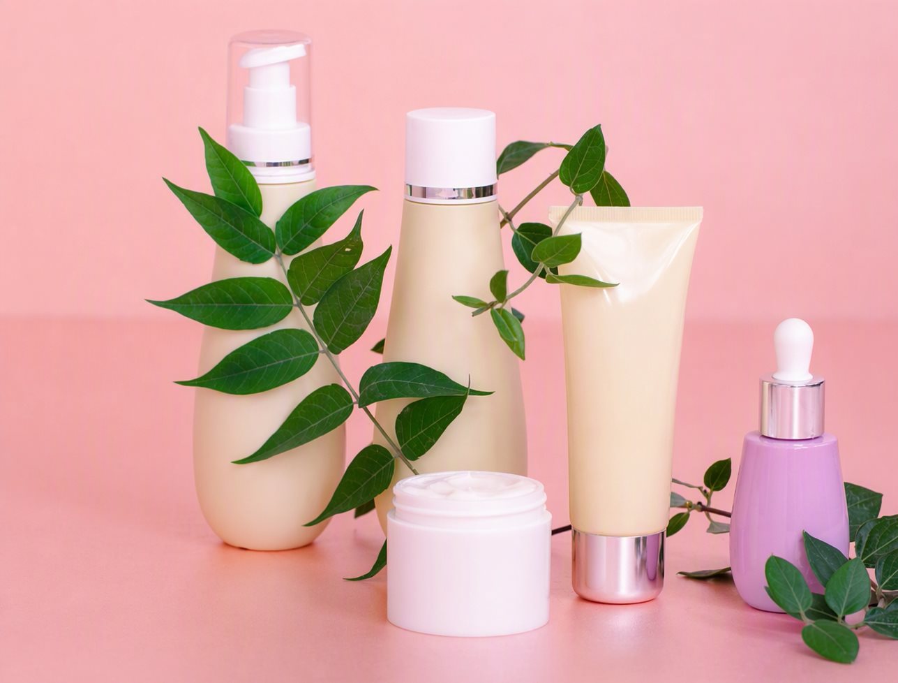 Five-piece skincare set: pump bottle, cream jar, squeeze tube and dropper bottle with green leaves on pink background